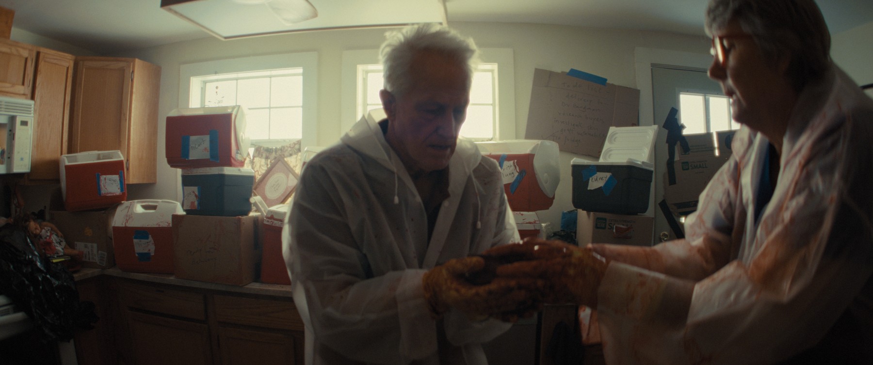 Elderly man holding a prosthetic organ in a kitchen full of labeled coolers