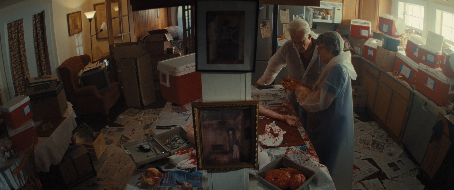 Elderly couple in a blood-spattered kitchen with a framed photograph